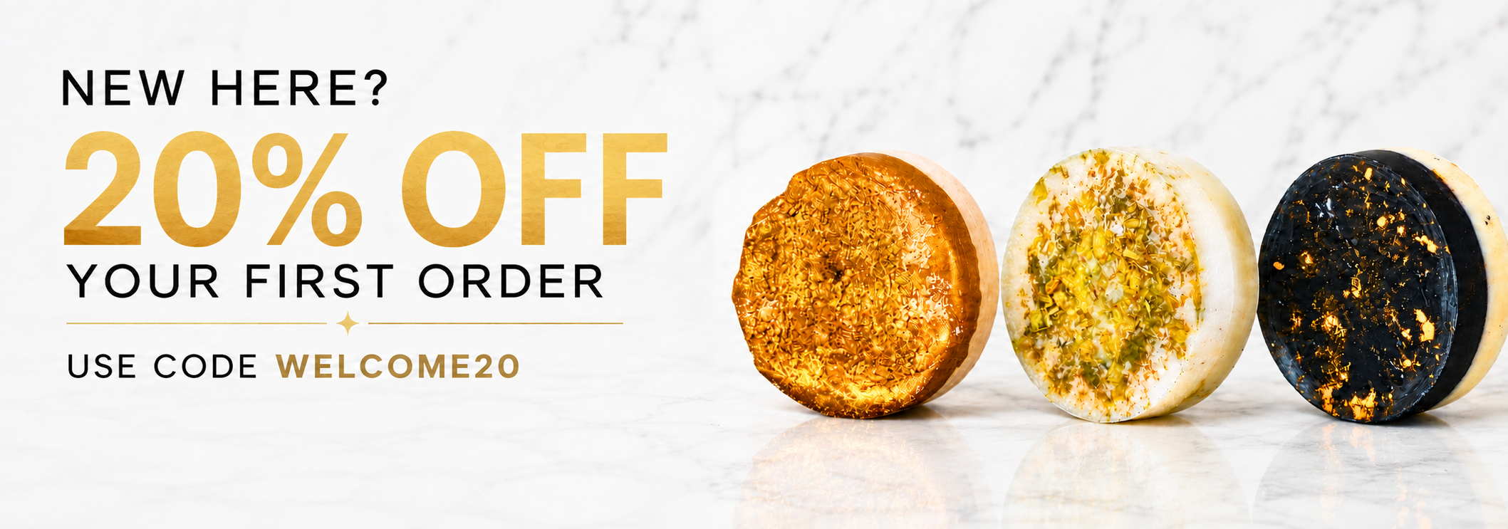 Promotional banner with text reading 'NEW HERE? 20% OFF YOUR FIRST ORDER' and 'USE CODE WELCOME20', alongside three round decorative soaps or candles on a light marble-like surface. The products have different finishes: one gold, one white with yellow speckles, and one black with gold flecks.